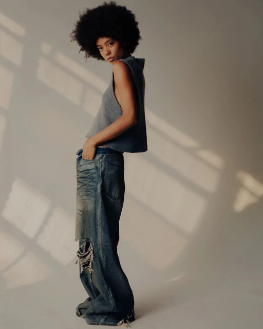 A young woman with afro-style hair poses in a photo studio, wearing a gray top and distressed, baggy jeans. Soft lighting creates shadows.