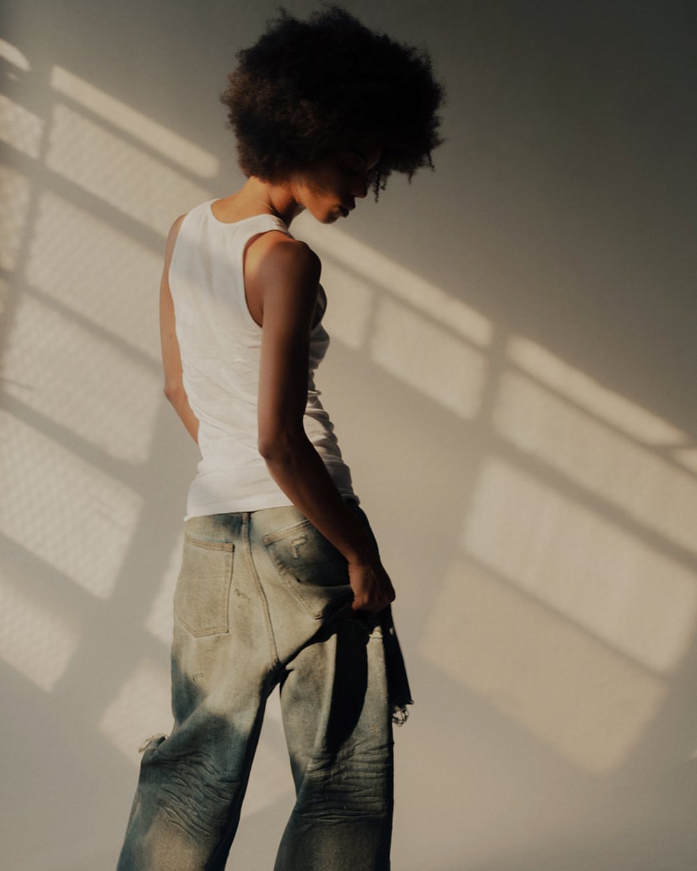 An adult with an afro hairstyle is captured in a medium shot, wearing a white tank top and distressed jeans, posing against a neutral wall.