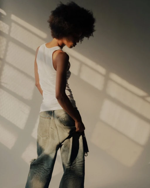 An adult with an afro hairstyle is captured in a medium shot, wearing a white tank top and distressed jeans, posing against a neutral wall.
