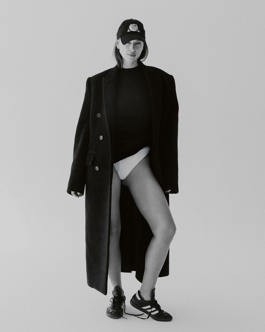 A woman in a baseball cap and overcoat poses in a black top and briefs, standing against a neutral backdrop in a fashion-forward portrait.