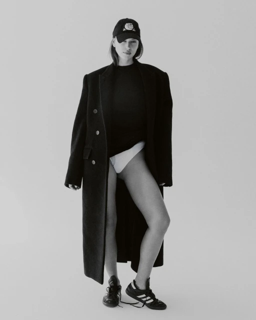 A woman in a baseball cap and overcoat poses in a black top and briefs, standing against a neutral backdrop in a fashion-forward portrait.