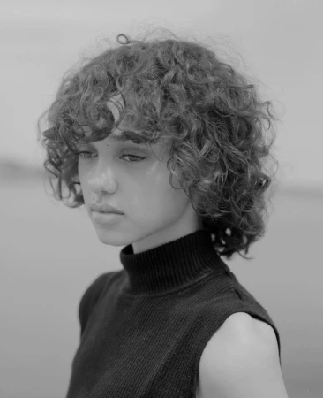Portrait of a young woman with curly short hair and a black turtleneck sweater against an out-of-focus background.