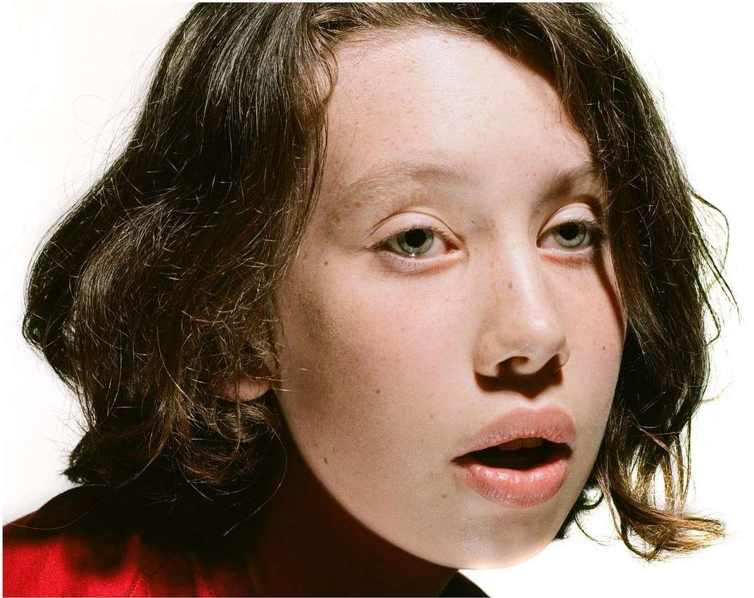 Close-up portrait of a young adult female with medium-length wavy brunette hair, light skin, and freckles against a bright white backdrop.
