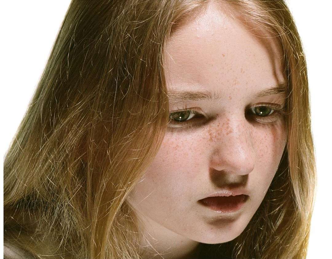 Close-up of a young woman with fair skin, freckles, and long, wavy blonde hair looking down against a plain white backdrop.