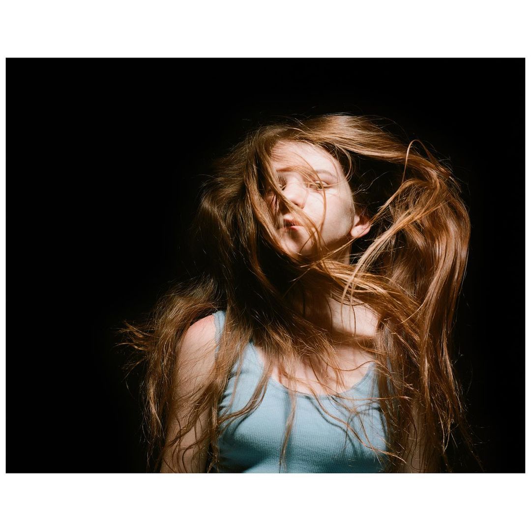 Portrait of a young woman with long, red hair. The shot is set against a dark background with studio lighting.