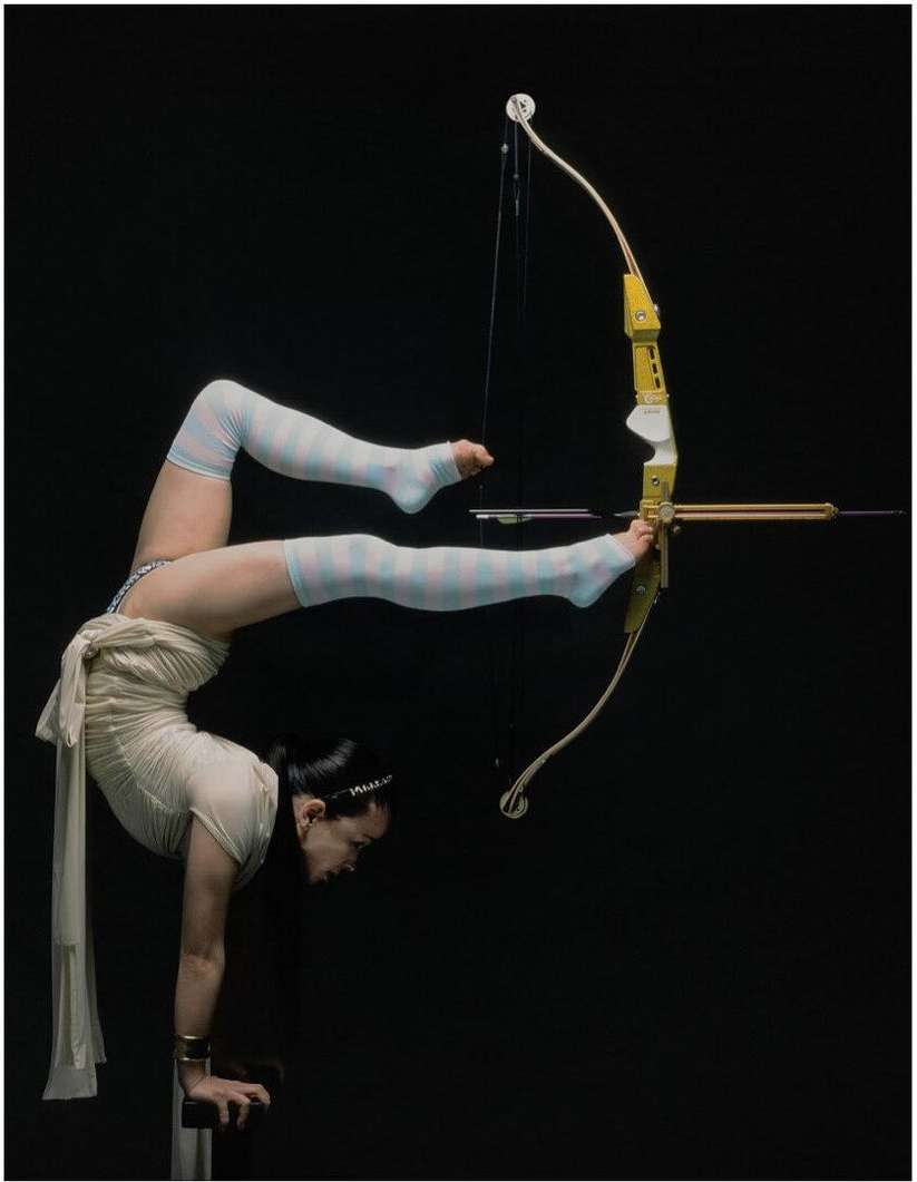 Adult female in leotard and striped thigh-highs performs acrobatic handstand with an archery bow in a dark indoor setting.