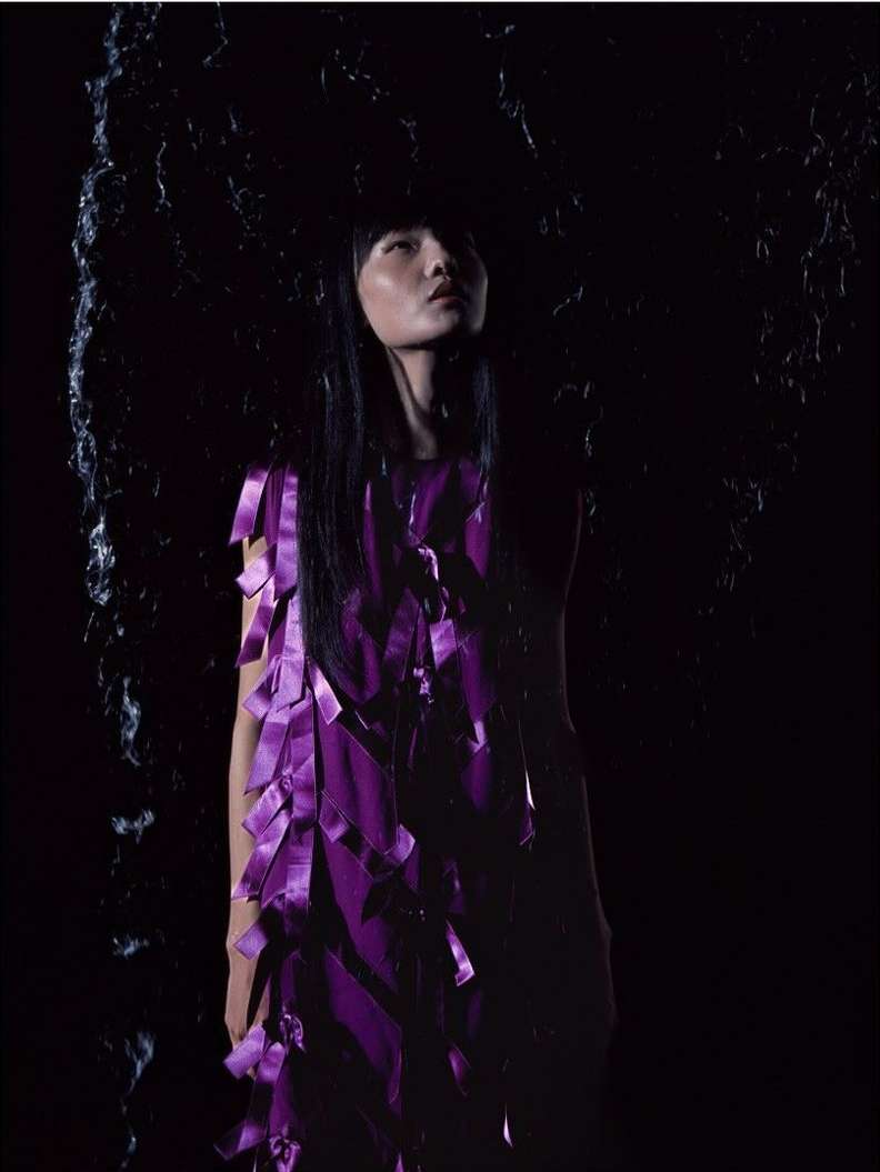 Asian woman in a purple dress with ribbons gazes upward in a studio setting with dramatic lighting and a dark textured background.