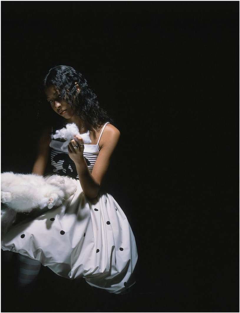 Young woman in a white dress with black polka dots holds a fluffy white object against a black background in a fashion style photo.