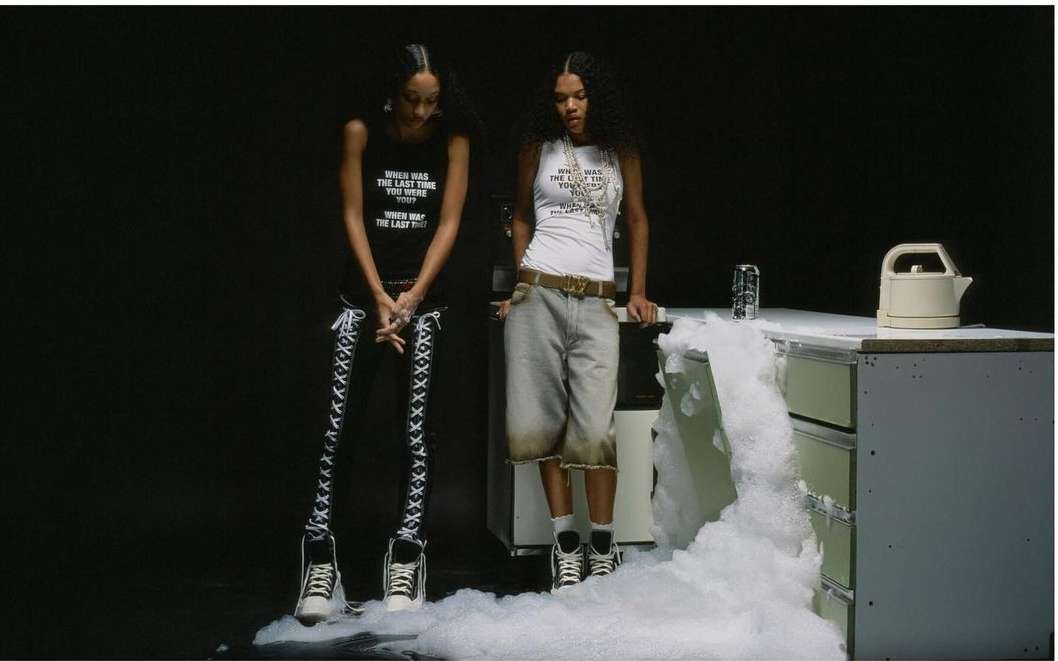 Two women in matching outfits stand near a foam-covered counter with a kettle and soda can against a dark background in a fashion shot.