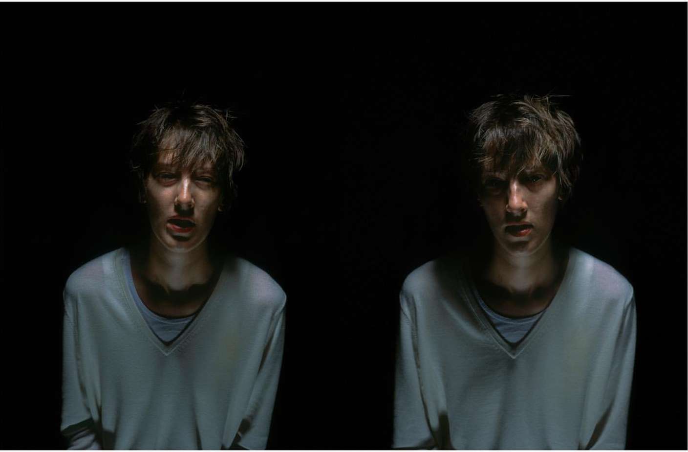 Two young adults in a dark indoor setting, with soft lighting emphasizing their faces and neutral-colored clothing, creating a dramatic effect.
