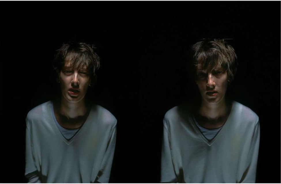Two young adults in a dark indoor setting, with soft lighting emphasizing their faces and neutral-colored clothing, creating a dramatic effect.