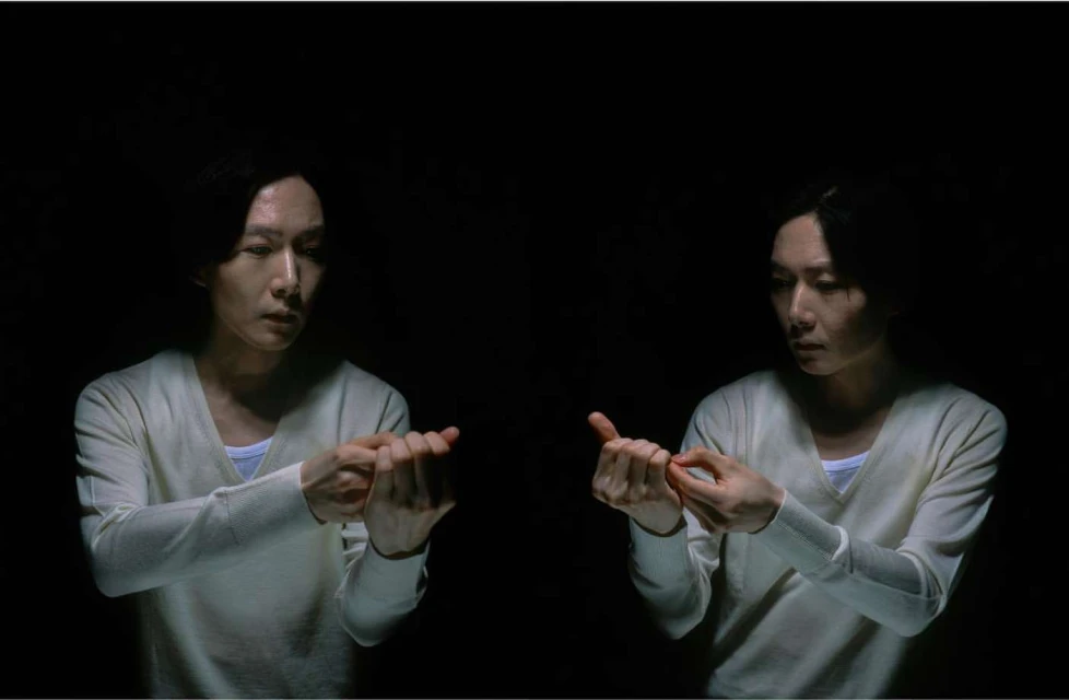 A split-screen effect with two Asian adults dressed in light sweaters, each holding their hands in front in an indoor setting with dark lighting.