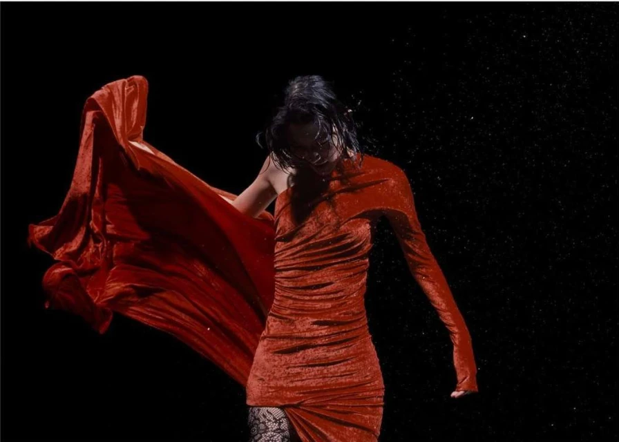 A woman in a red dress poses dramatically in a dark studio, fabric billowing, creating a striking fashion portrait.