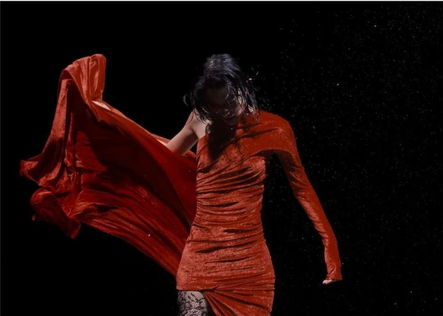 A woman in a red dress poses dramatically in a dark studio, fabric billowing, creating a striking fashion portrait.