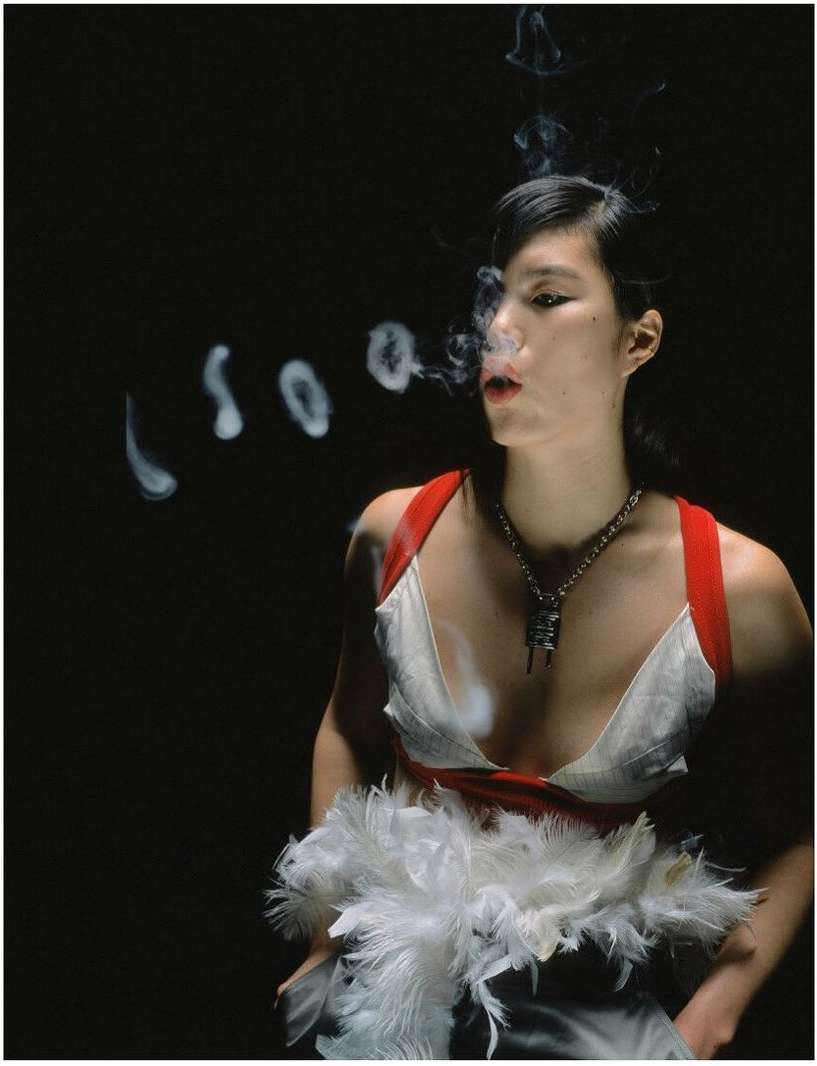 Woman exhales smoke rings in a dimly lit space, wearing a low-cut top with red straps and white feathers. A silver necklace is visible.
