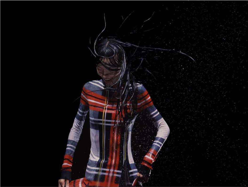 Fashion shot of a woman with wet hair wearing a red plaid dress in a dark studio with water droplets visible against a black backdrop.