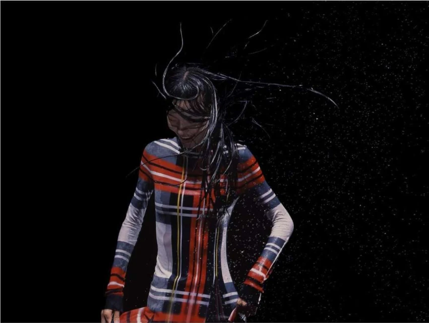 Fashion shot of a woman with wet hair wearing a red plaid dress in a dark studio with water droplets visible against a black backdrop.