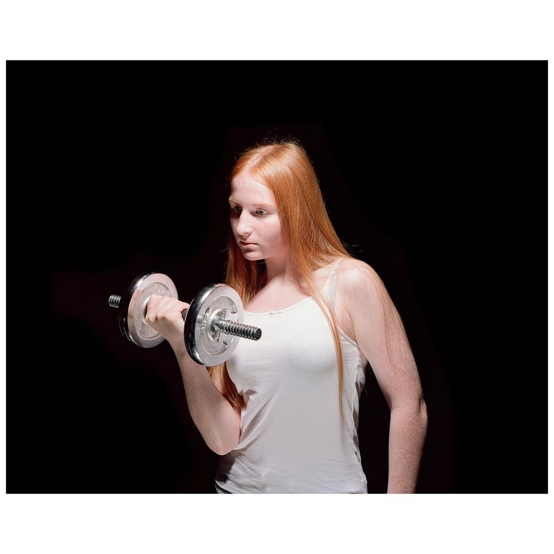 A woman with red hair works out with a silver dumbbell in a studio setting against a dark background, highlighting her strength and focus.