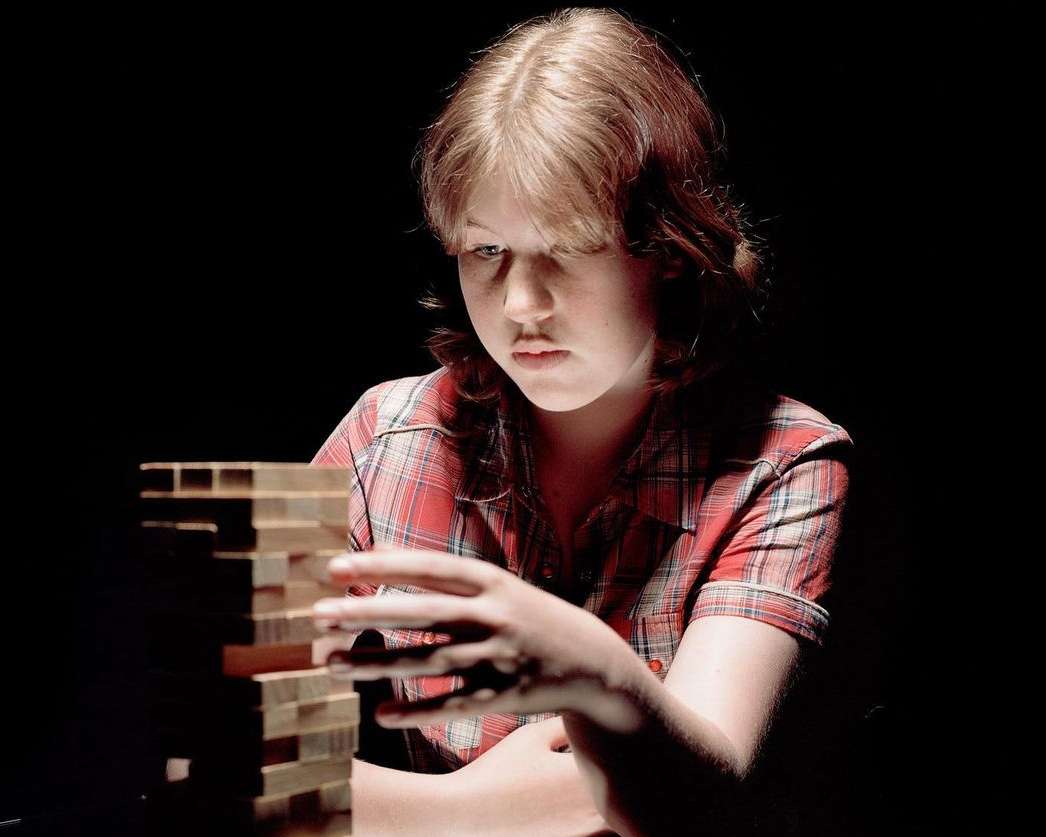 Young adult woman in red plaid shirt concentrates on removing wood block from partially constructed tower on a dark background.