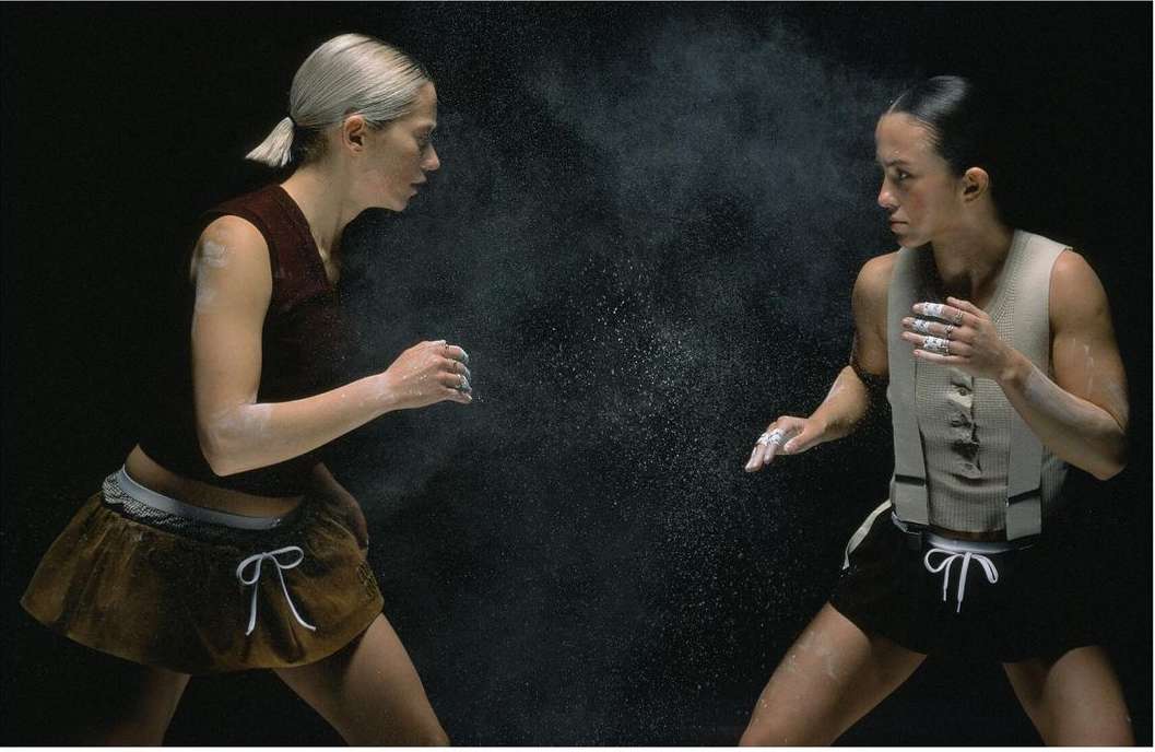 Two women in athletic wear facing each other, with a burst of chalk dust between them, set against a stark black background.