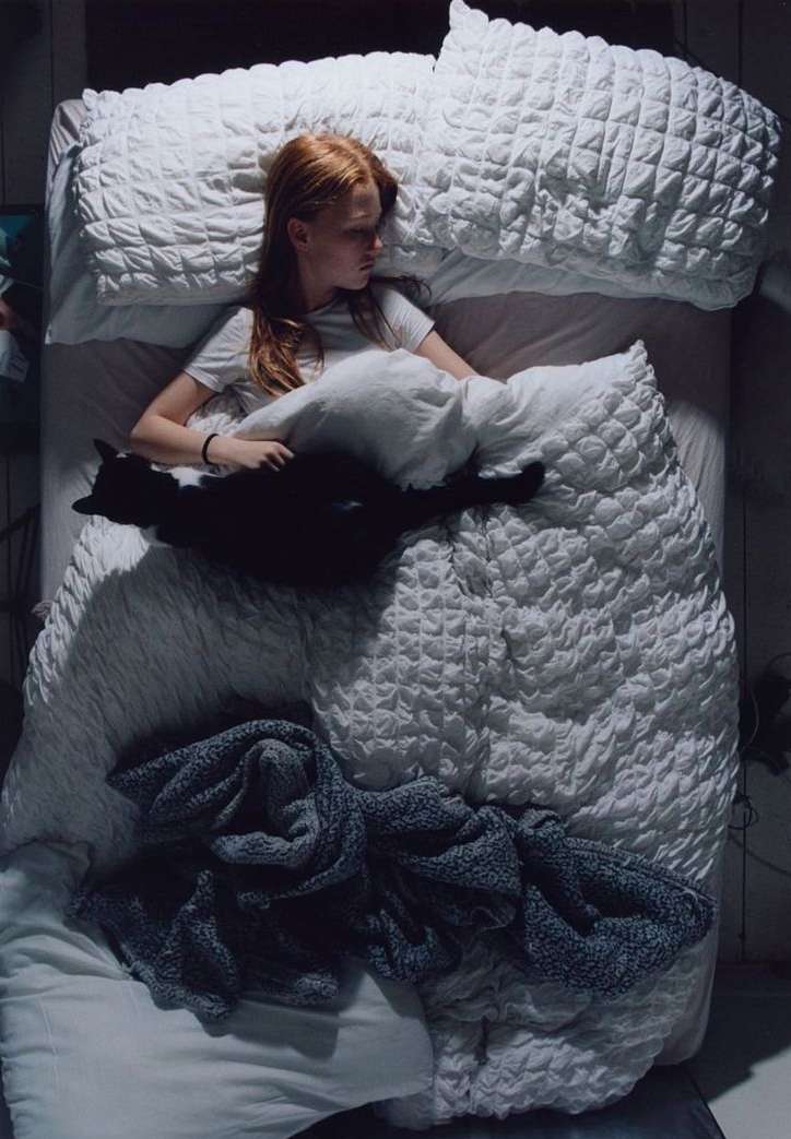 Red-haired person sleeps in a white bed with a black cat curled up next to them, covered by a white down comforter and soft blanket.
