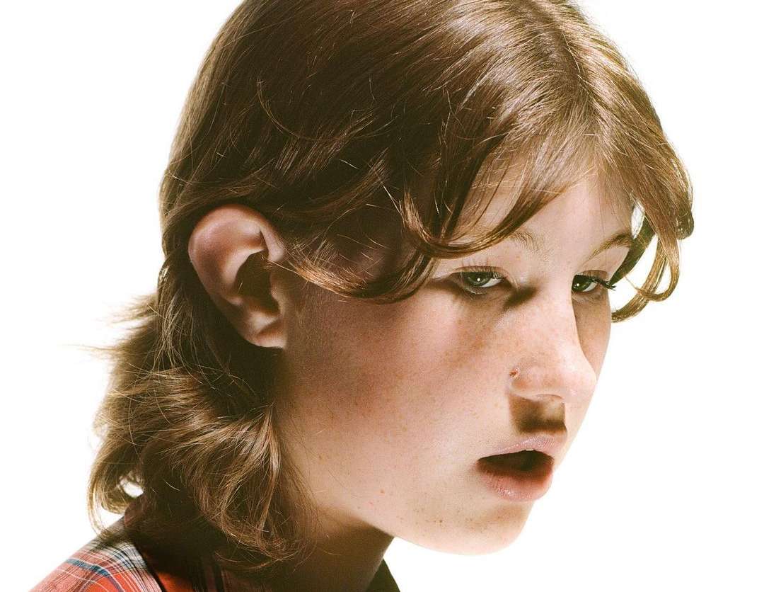 Close-up portrait of a young person with brown hair and fair skin, wearing a red plaid top, against a white background.