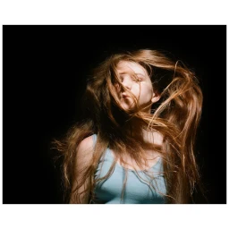 Close-up portrait of a young woman with long brown hair whipping across her face against a black background