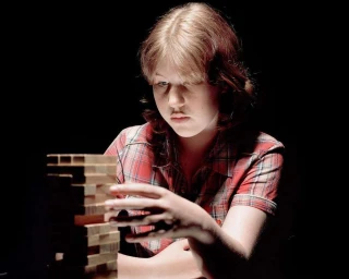 Young woman in a red plaid shirt focusing intently as she plays a wooden tower game against a dark background