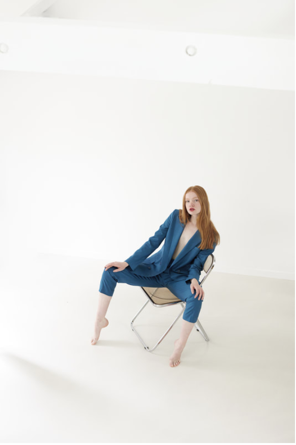 Red-haired woman in blue suit poses barefoot on silver folding chair in bright photo studio with white background and natural lighting.