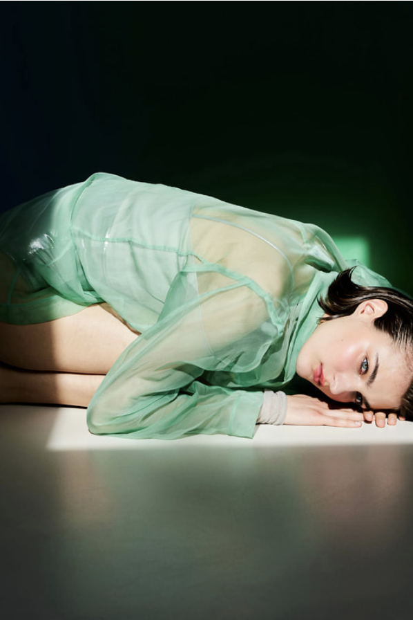 Woman in sheer green top laying on a white surface, head resting on her hands. Dark background and soft lighting.