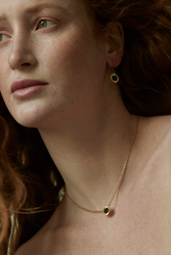 Close-up of a fair-skinned woman with red hair wearing gold jewelry: earrings and a necklace with a circular pendant.