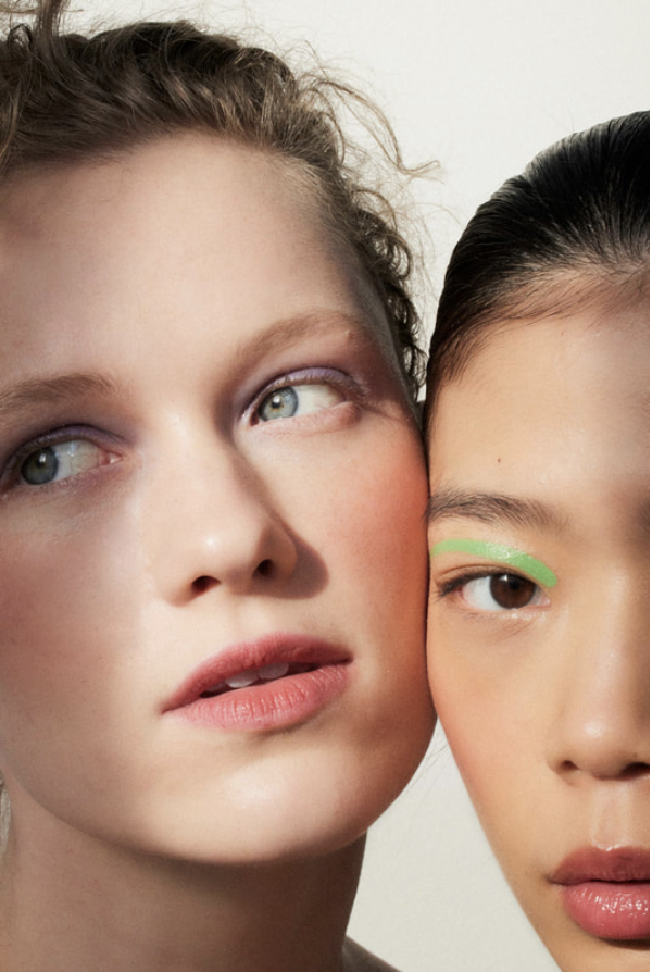 Close-up portrait of two women with creative makeup, one with a light skin tone and purple eyeshadow, the other with a dark skin tone and green eyeliner.