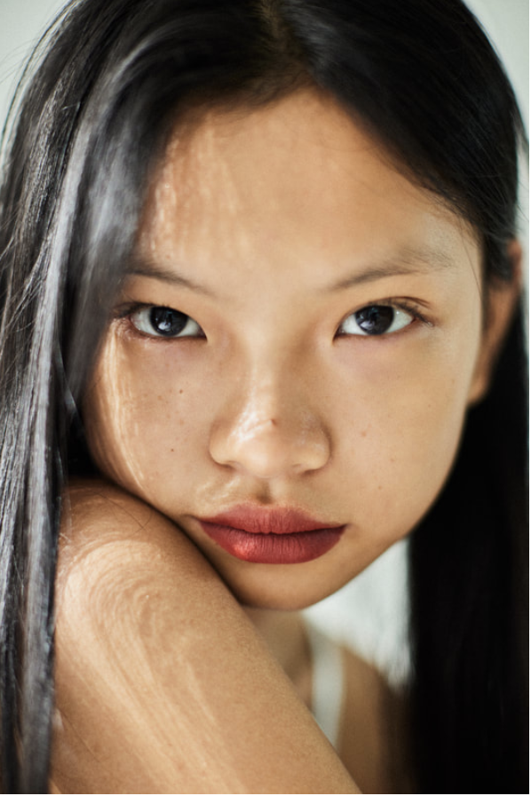 Close-up portrait of a young Asian woman with long dark hair, fair skin, and red lipstick, looking directly at the camera in a studio setting.