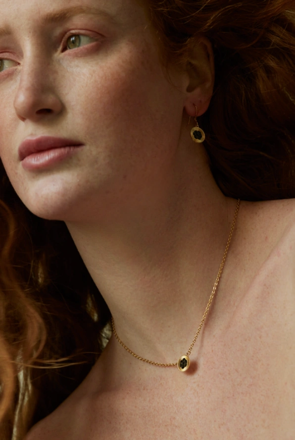 Close-up portrait of a red-haired woman with freckles wearing a delicate gold chain necklace with a pendant and gold earring.
