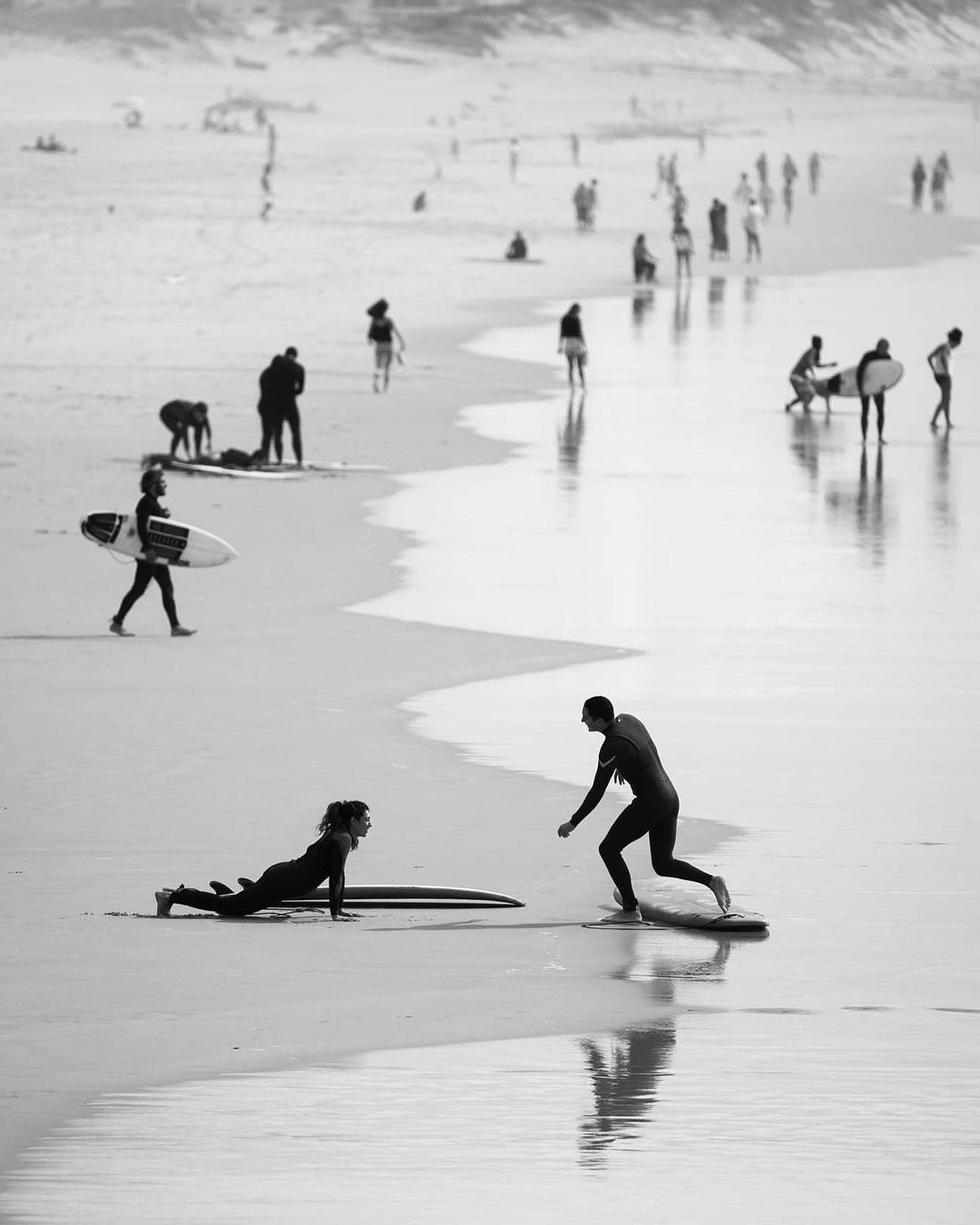 Black and white shot of people on a beach; a man teaches a woman to surf while others walk along the water's edge in the background.