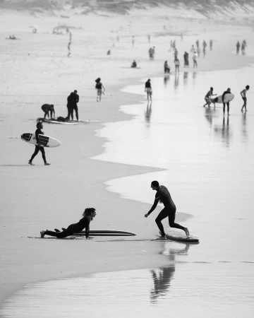 Black and white shot of people on a beach; a man teaches a woman to surf while others walk along the water's edge in the background.