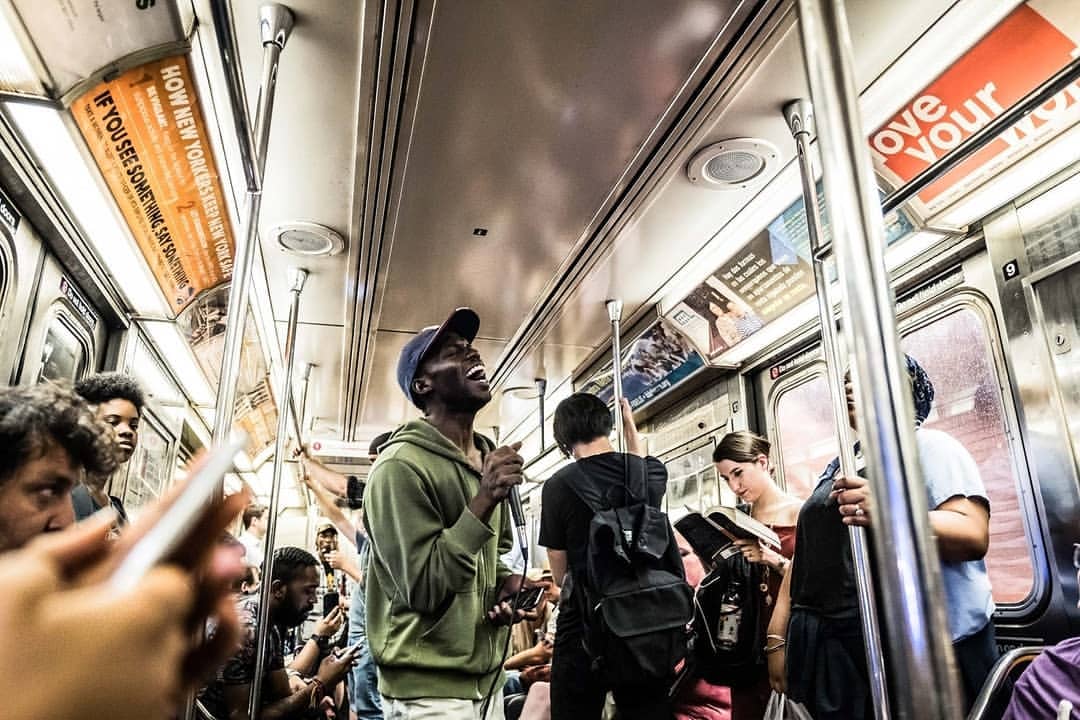 Man singing on a subway train, surrounded by other passengers. Subway interior with advertisements visible in the background.