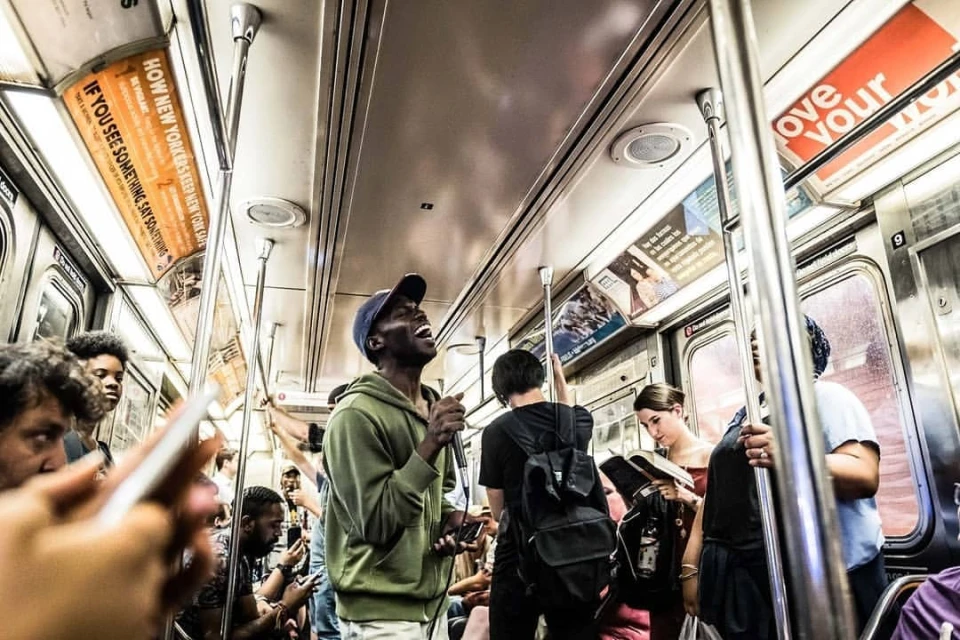 Man singing on a subway train, surrounded by other passengers. Subway interior with advertisements visible in the background.