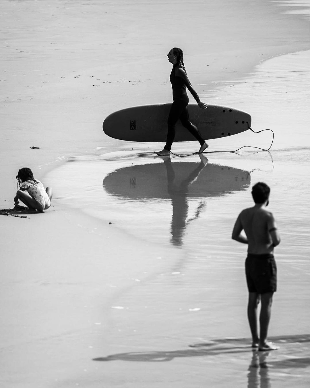Monochrome beach scene with a woman carrying a surfboard, a kneeling child, and a standing man.