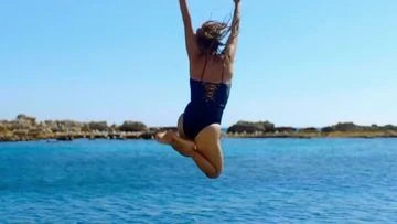 Woman in a black swimsuit leaps joyfully over the blue sea against a backdrop of clear skies and rocky formations on the horizon.