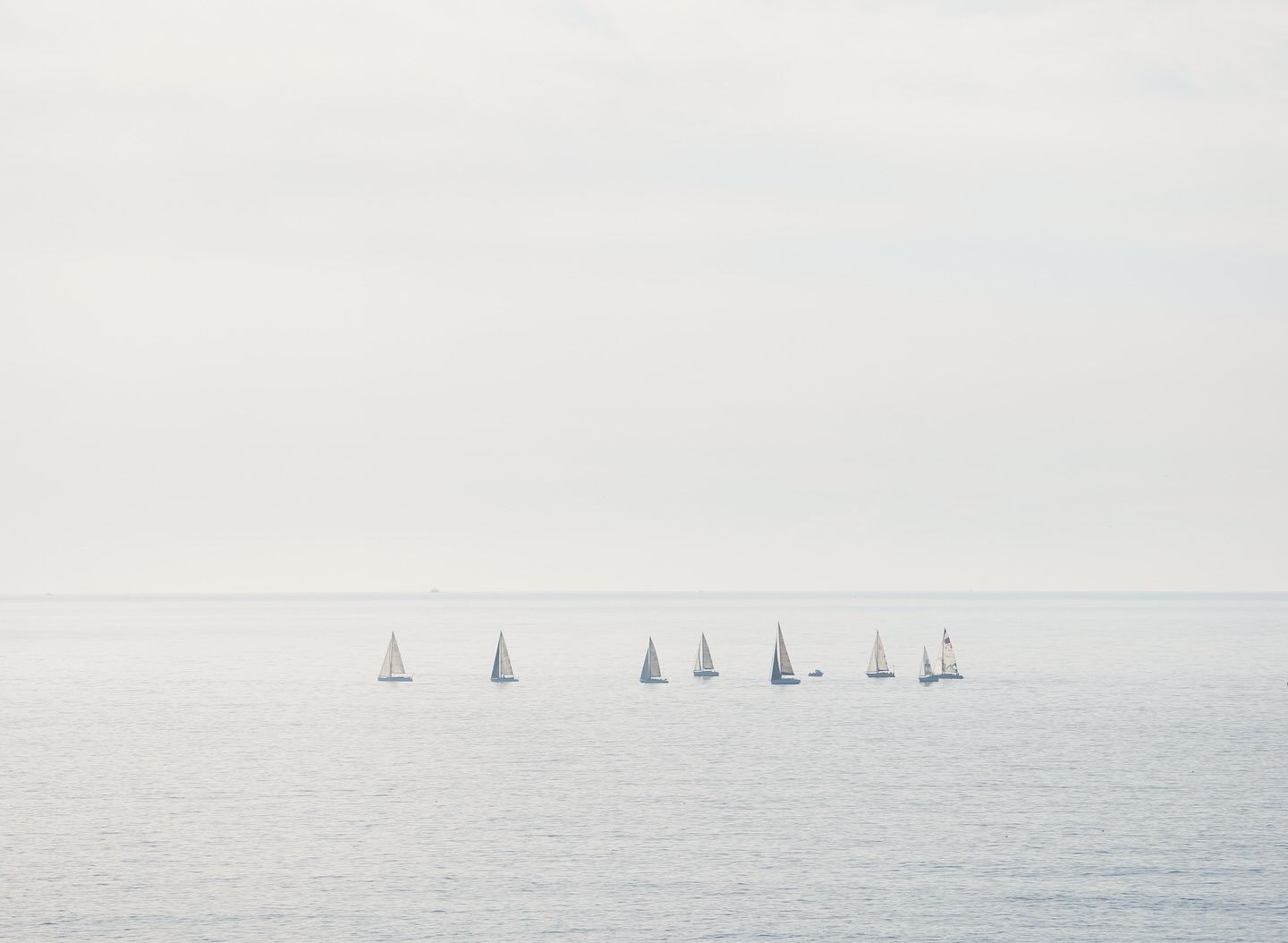 A group of sailboats are sailing on the water in a hazy, overcast day. The composition has a soft, muted color palette.