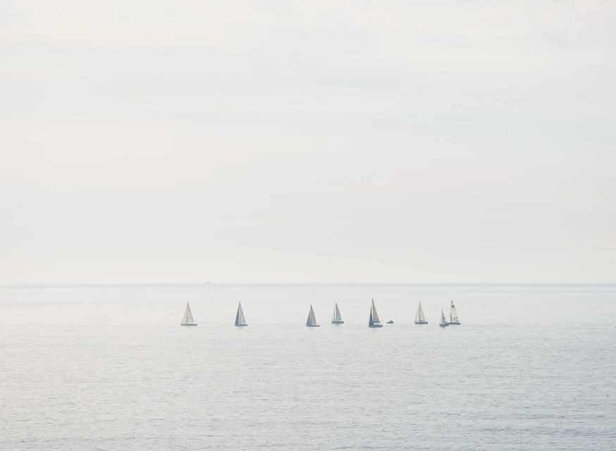 A group of sailboats are sailing on the water in a hazy, overcast day. The composition has a soft, muted color palette.