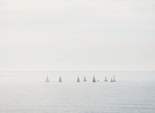 A group of sailboats are sailing on the water in a hazy, overcast day. The composition has a soft, muted color palette.