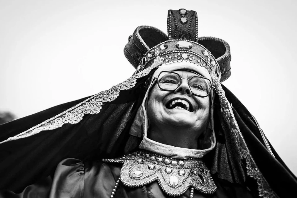 Smiling woman in elaborate crown and jeweled collar, portrait in black and white, looking upward with joyous expression on her face.