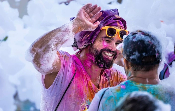 Smiling man wearing yellow sunglasses and a purple head covering covered in colourful foam and powder
