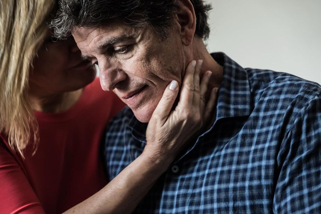 Mature man comforted by woman with hands on his face in a close up studio shot, the man looks sad in a blue plaid shirt.