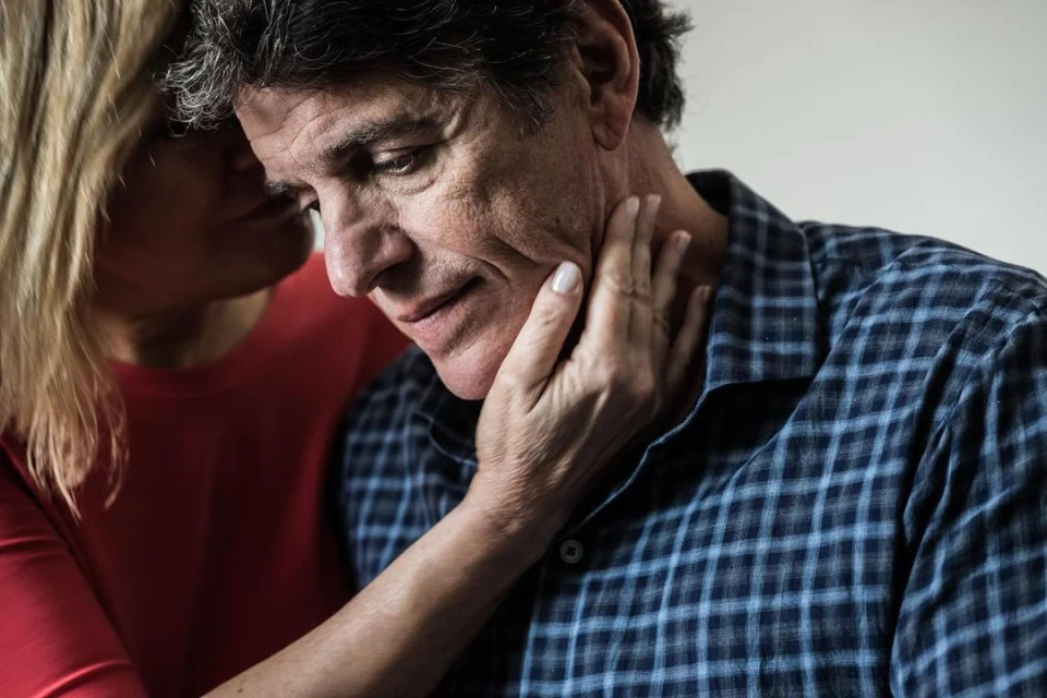 Mature man comforted by woman with hands on his face in a close up studio shot, the man looks sad in a blue plaid shirt.