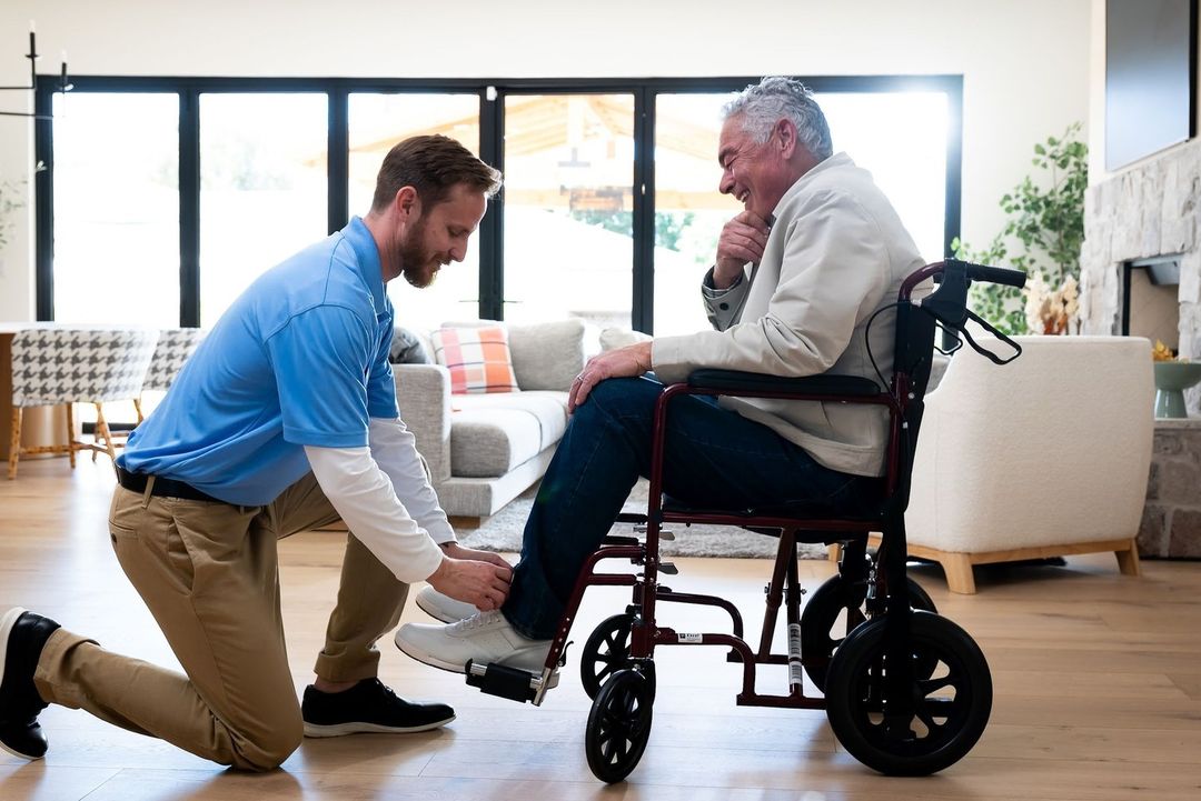 Healthcare scene: An adult male healthcare worker assists an elderly man in a wheelchair, indoors, in a home setting with natural light.