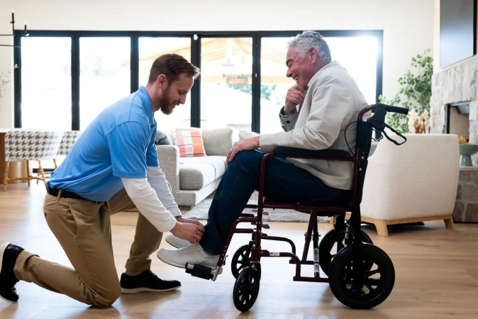 Healthcare scene: An adult male healthcare worker assists an elderly man in a wheelchair, indoors, in a home setting with natural light.