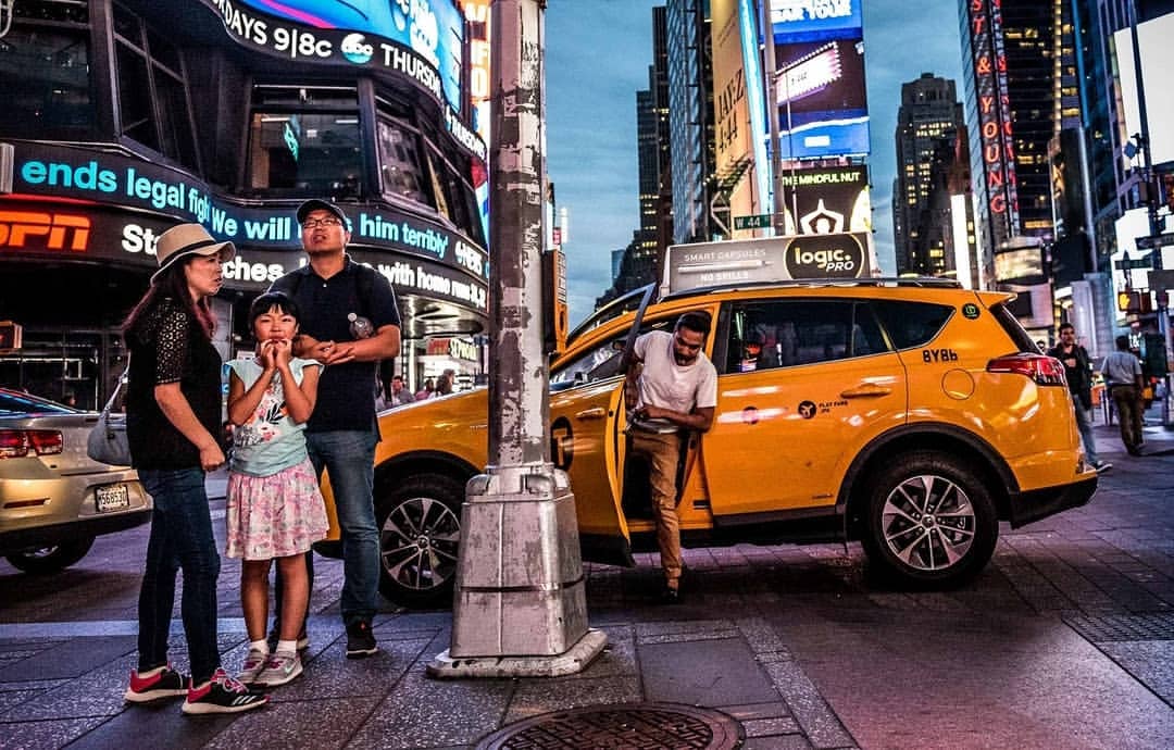 Family on a sidewalk in Times Square, New York City with a yellow taxi cab and illuminated billboards at dusk in an urban environment.